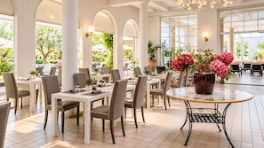 A spacious indoor dining area with multiple tables and chairs, large windows allowing natural light, and potted plants adding greenery. - Bellavista Terme Resort & Spa (Montegrotto Terme)