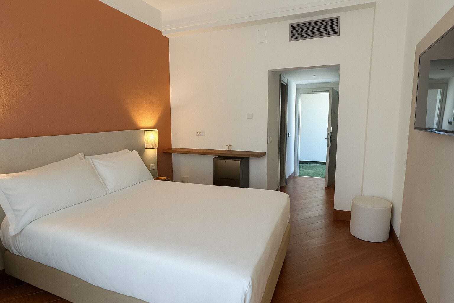 Quarto de hotel com uma cama grande, uma mesa de cabeceira, um abajur e uma pequena lixeira.