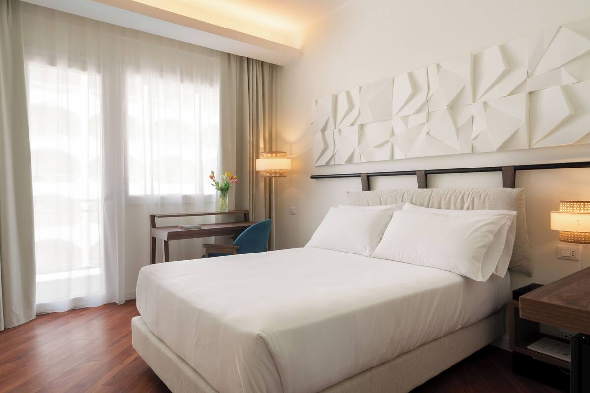 Une chambre d’hôtel avec un grand lit, un bureau avec une chaise, une lampe et une œuvre d’art abstraite accrochée au mur.