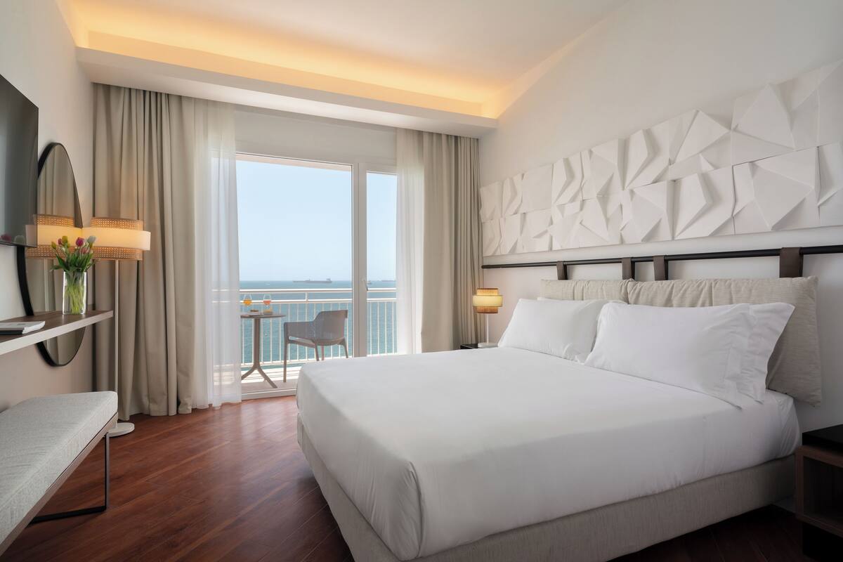 Une chambre d’hôtel moderne avec un grand lit, un balcon offrant une belle vue et un panneau décoratif mural.