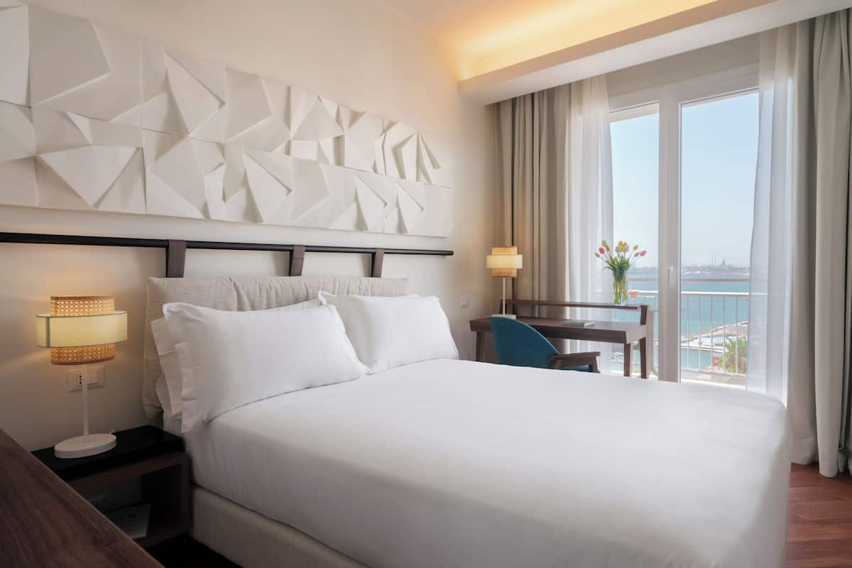 Une chambre d’hôtel avec un grand lit, un bureau avec une chaise, une vue sur la mer et un panneau décoratif au mur.
