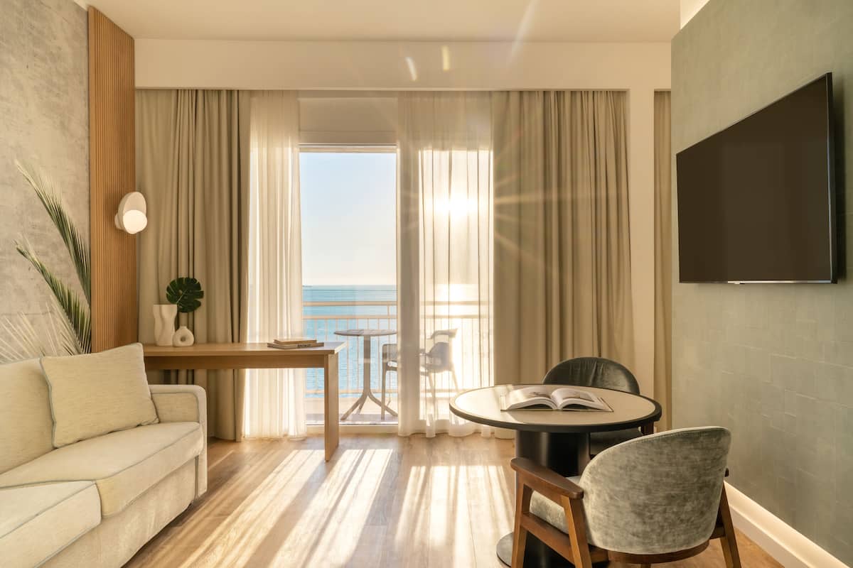 Une chambre d’hôtel moderne équipée d’un canapé, d’une table ronde, d’une chaise, d’un téléviseur et d’un balcon avec vue sur la mer.