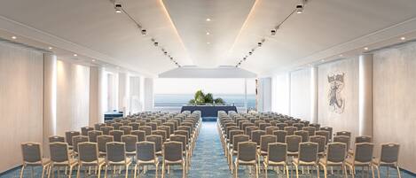 Un salón de conferencias con hileras de sillas mirando hacia un escenario, ventanas amplias y vistas al océano.