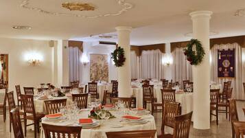 A formal dining room with multiple tables set for service, elegant chairs, and decorative wreaths.