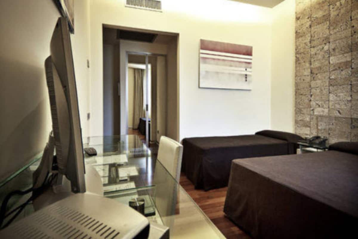 A hotel room with a glass desk, two beds, a computer, and a brick wall.
