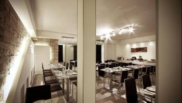 A modern dining area with long tables and chairs, a brick wall on one side, and a kitchen area visible in the background.