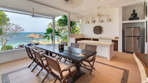 A modern kitchen with a central island, a dining area with wooden chairs, and a view of the ocean. - Napasai Samui (Koh Samui)