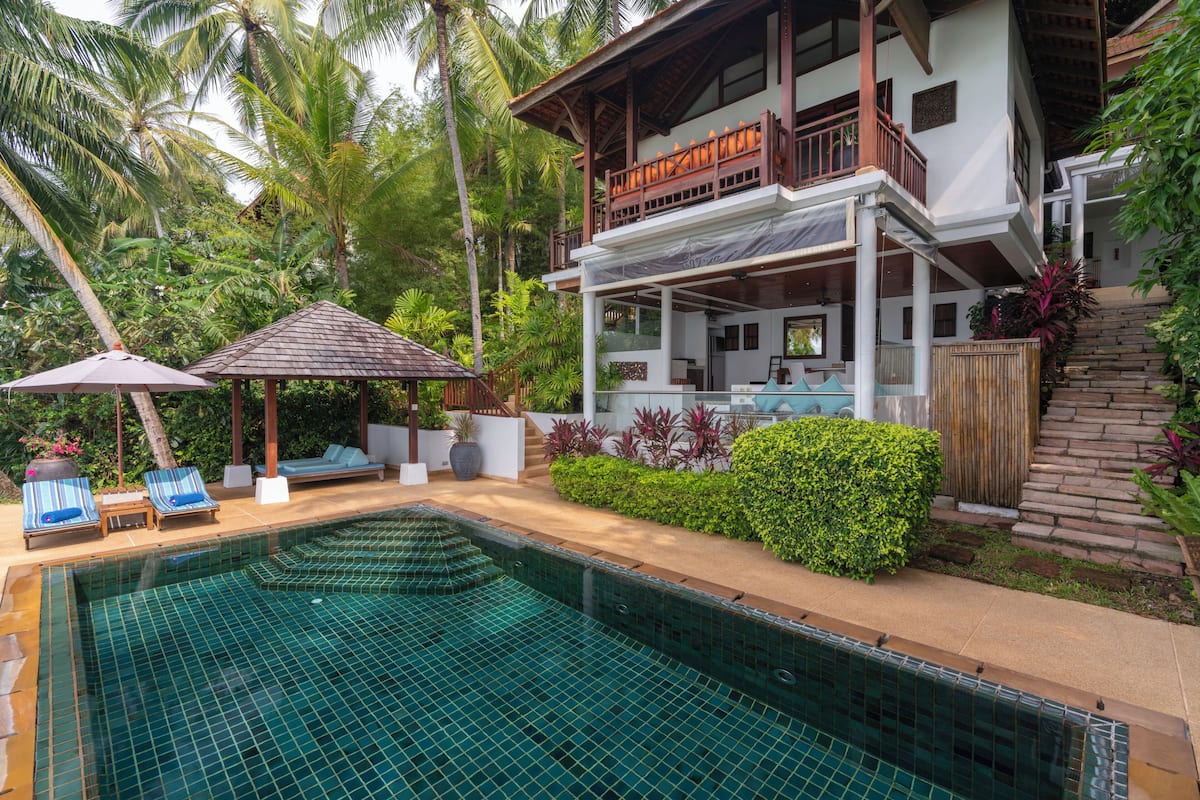 A pool area with lounge chairs, a covered seating area, and a two-story building with a balcony.