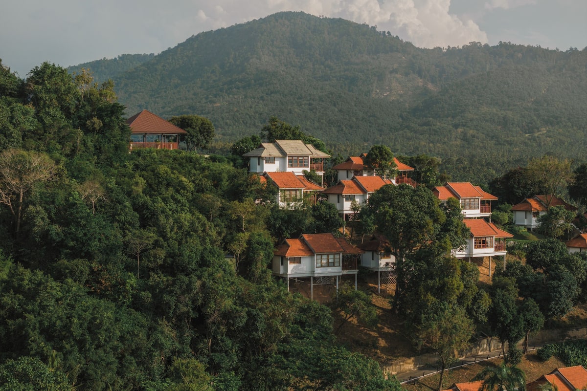 A hillside residential area with houses, some with balconies, surrounded by dense greenery and a mountain in the background.