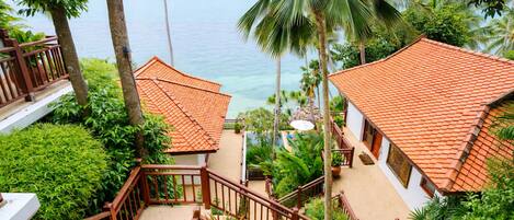 A coastal hotel with a staircase leading to a balcony overlooking the sea, surrounded by lush greenery and palm trees.