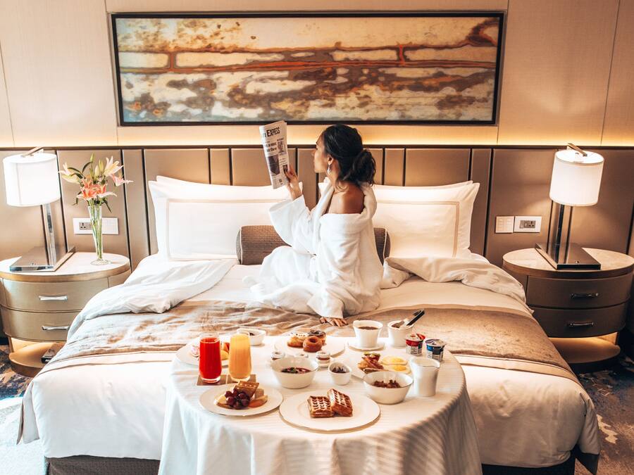 A hotel room with a bed, a breakfast table with food and drinks, a lamp, and a painting on the wall.