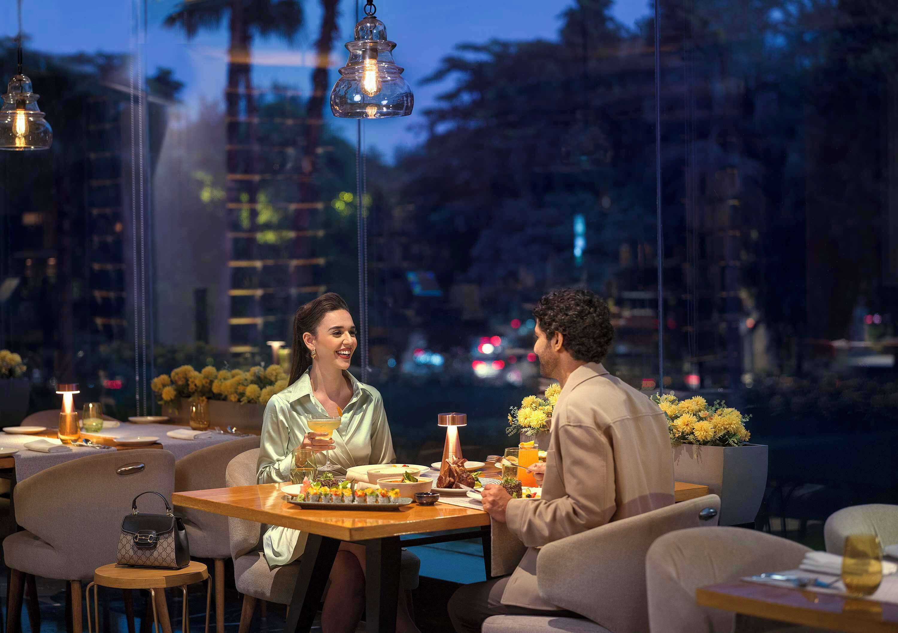 A couple dining at a table with a city view, a glass table, and ambient lighting.