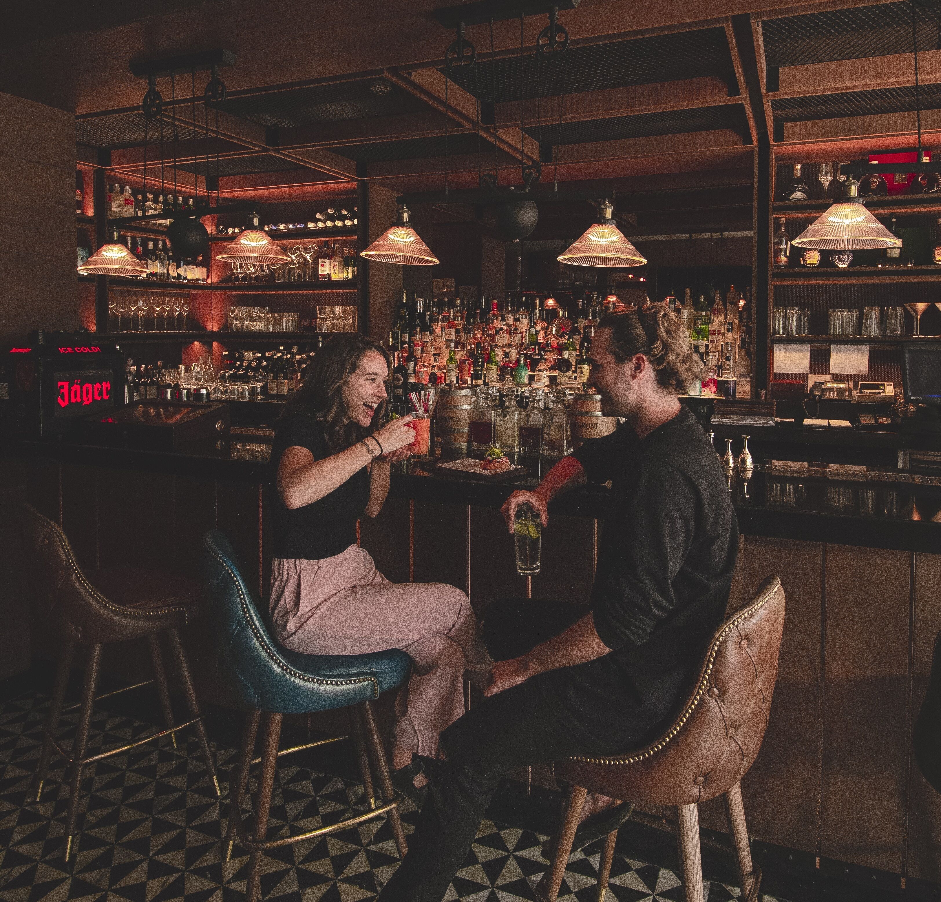 A bar with two stools, a person holding a drink, and shelves stocked with bottles and glasses.