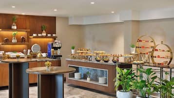 Zona de buffet con diferentes opciones de comida, una máquina de café y plantas decorativas.
