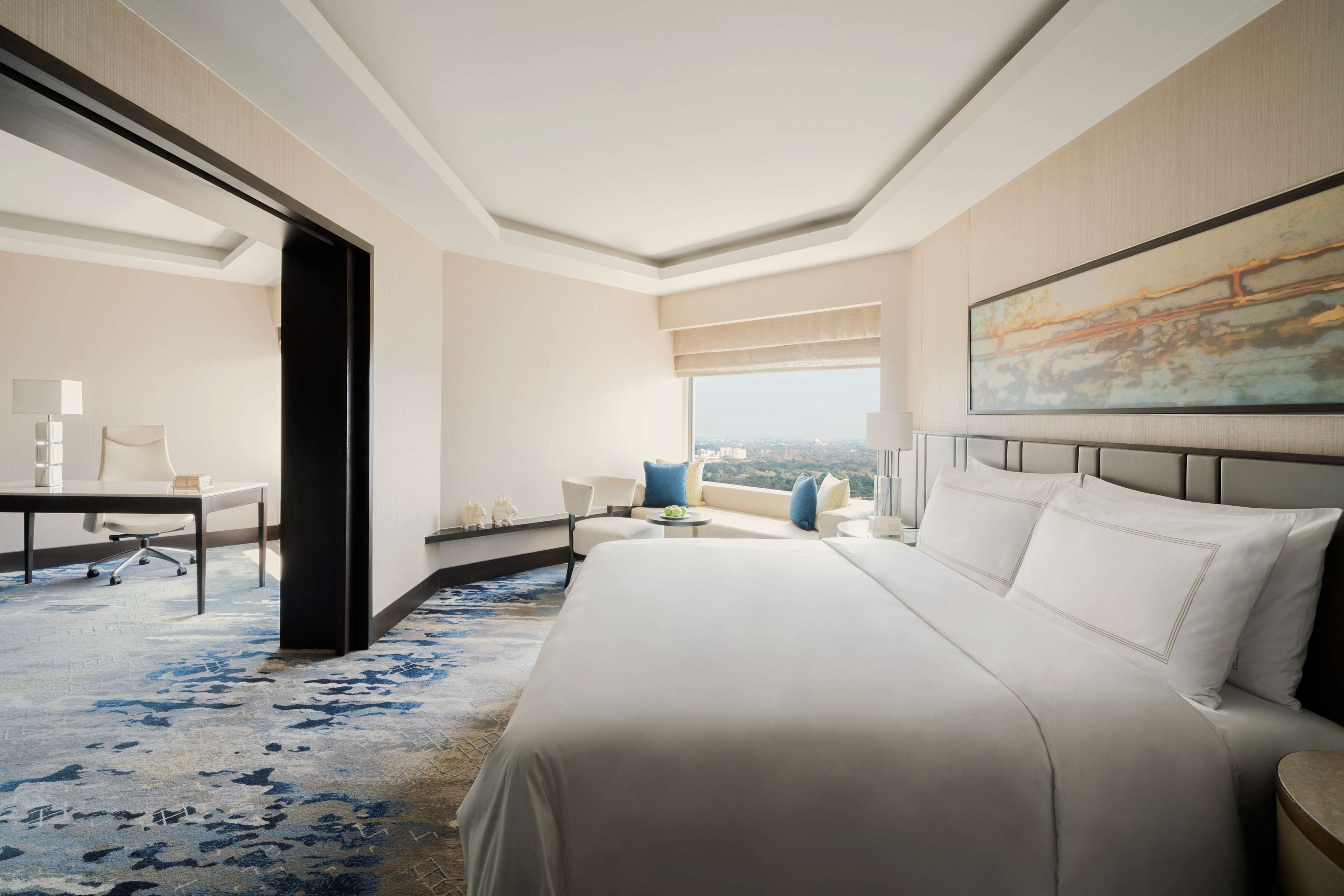 A hotel room with a large bed, a desk, a chair, and a sofa. There is a window with a view of the city.