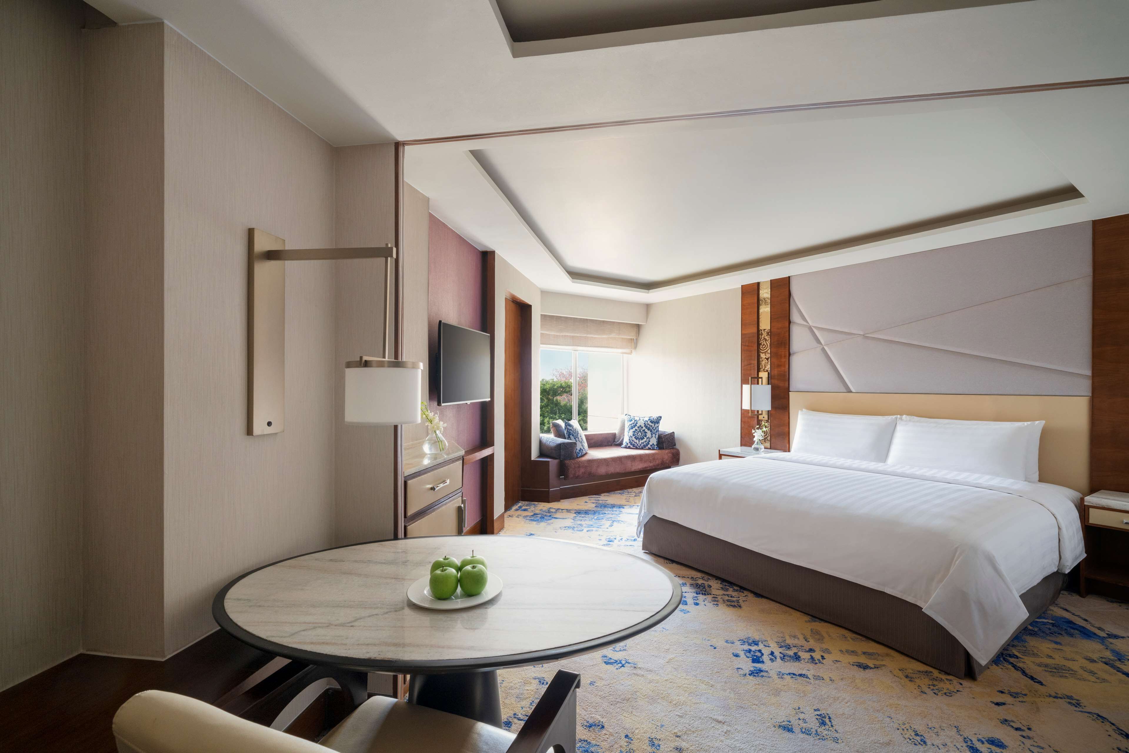 A hotel room with a large bed, a round dining table with apples, a TV, and a view of the outdoors.