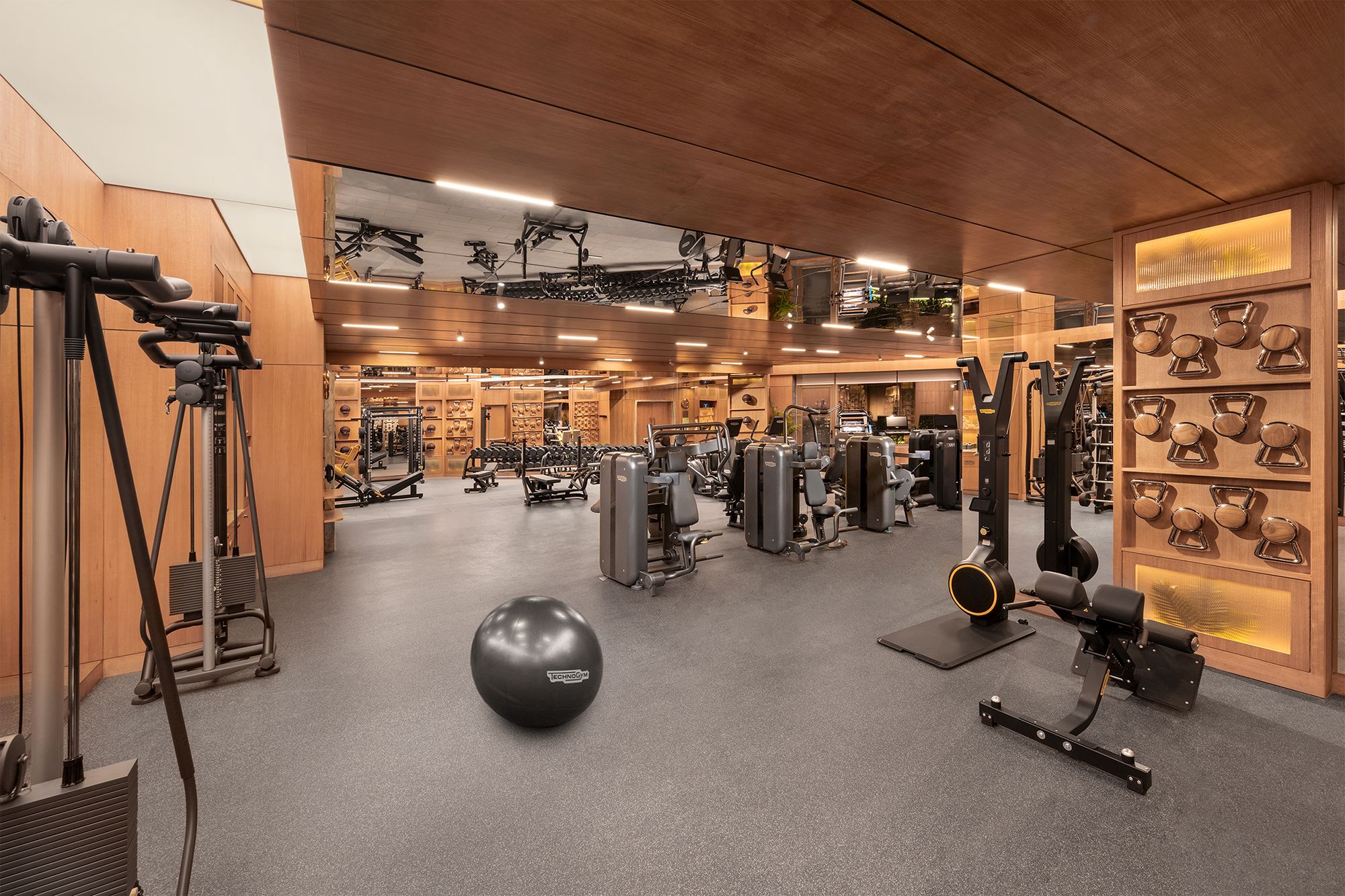 Une salle de sport équipée de divers appareils d’exercice, dotée d’un mur avec des miroirs et d’un autre mur où sont rangés des kettlebells.