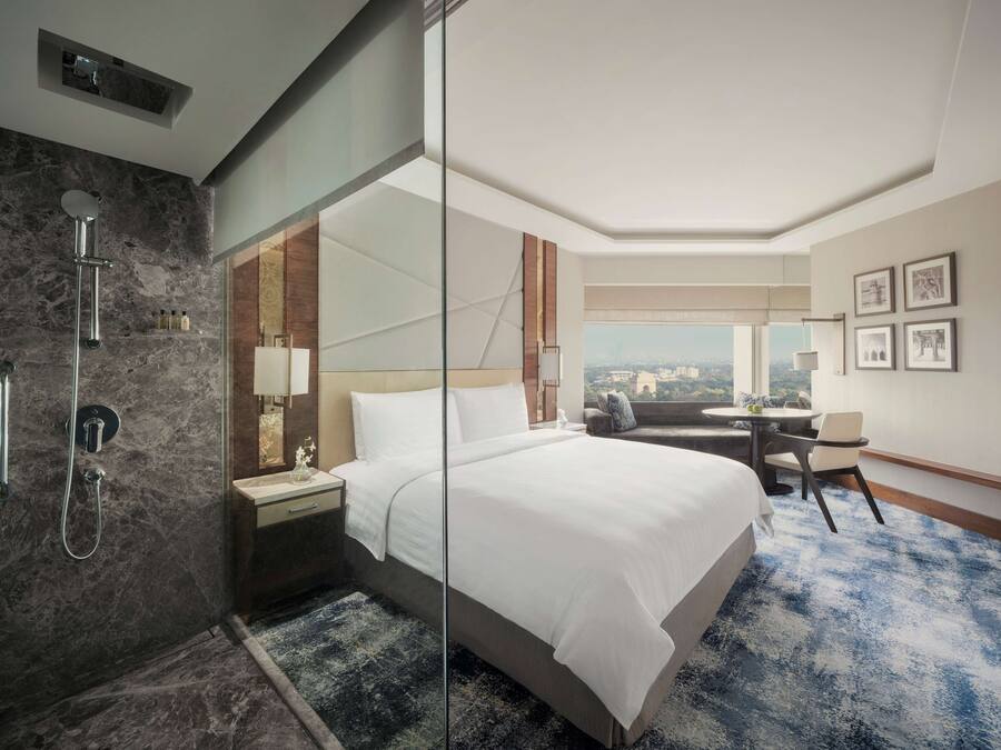 A hotel room with a large bed, a glass shower enclosure, a small desk with a chair, and a view of the city.