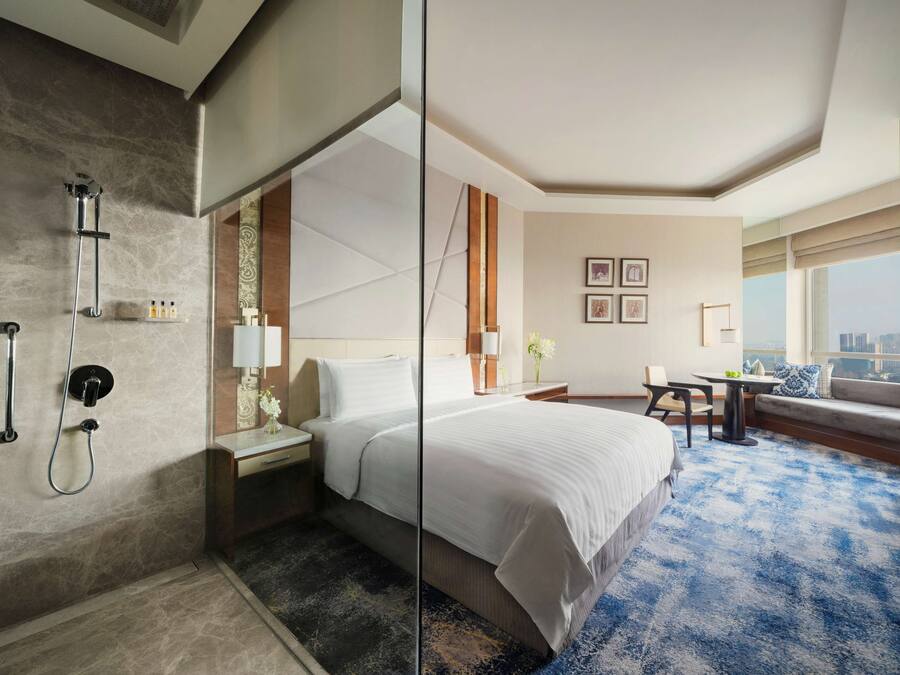 A hotel room with a large bed, a desk, a chair, a window with a city view, and a shower area.