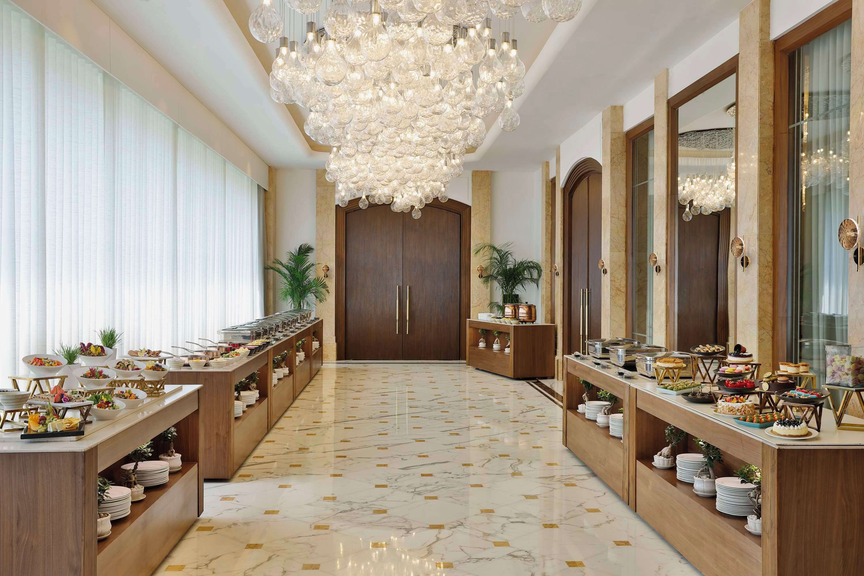 Amplia zona de buffet con suelo de mármol y mostradores de madera, que ofrece una variedad de alimentos y una gran lámpara de araña.