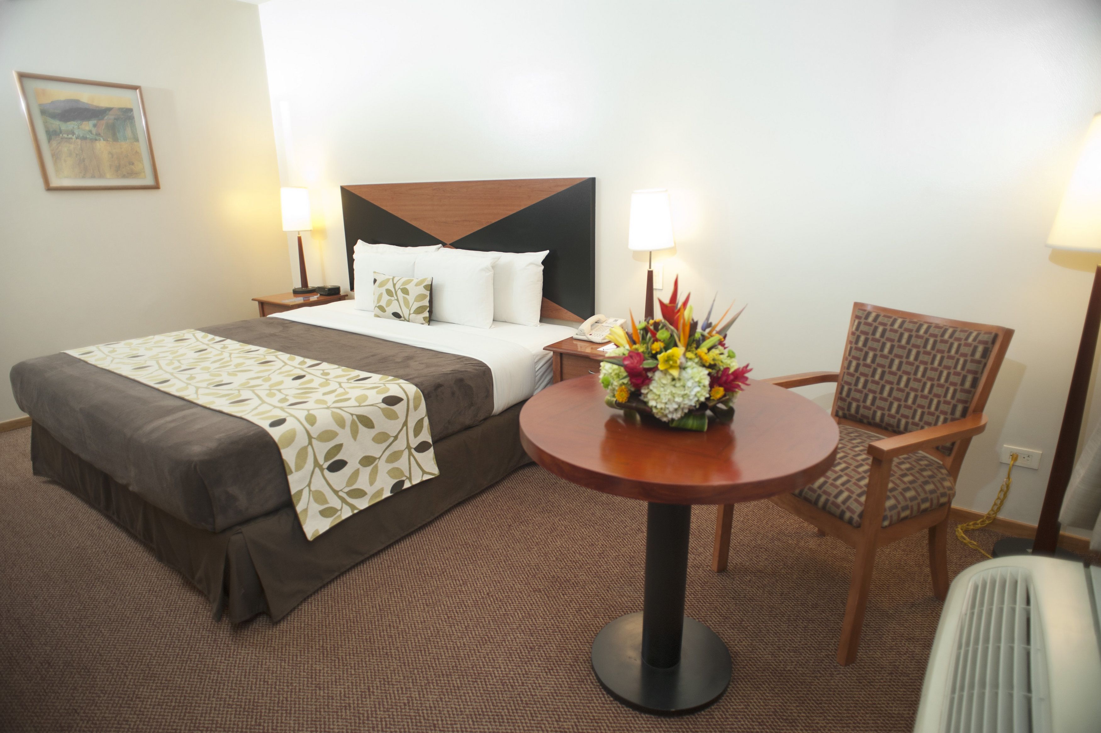 Une chambre d’hôtel avec un lit, une table ronde agrémentée d’un arrangement floral, une chaise et des lampes de chevet.