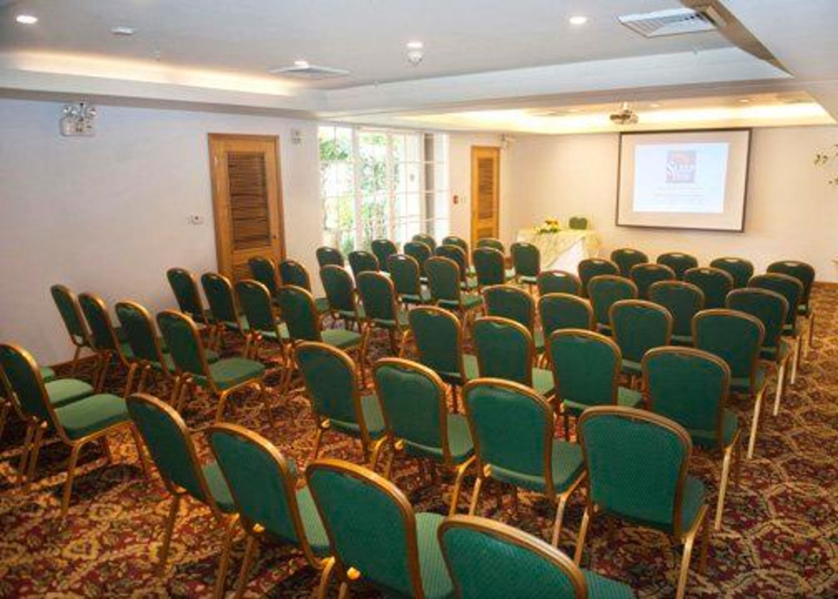 Un salón de conferencias con filas de sillas verdes, piso alfombrado, una pantalla de proyector y una puerta.