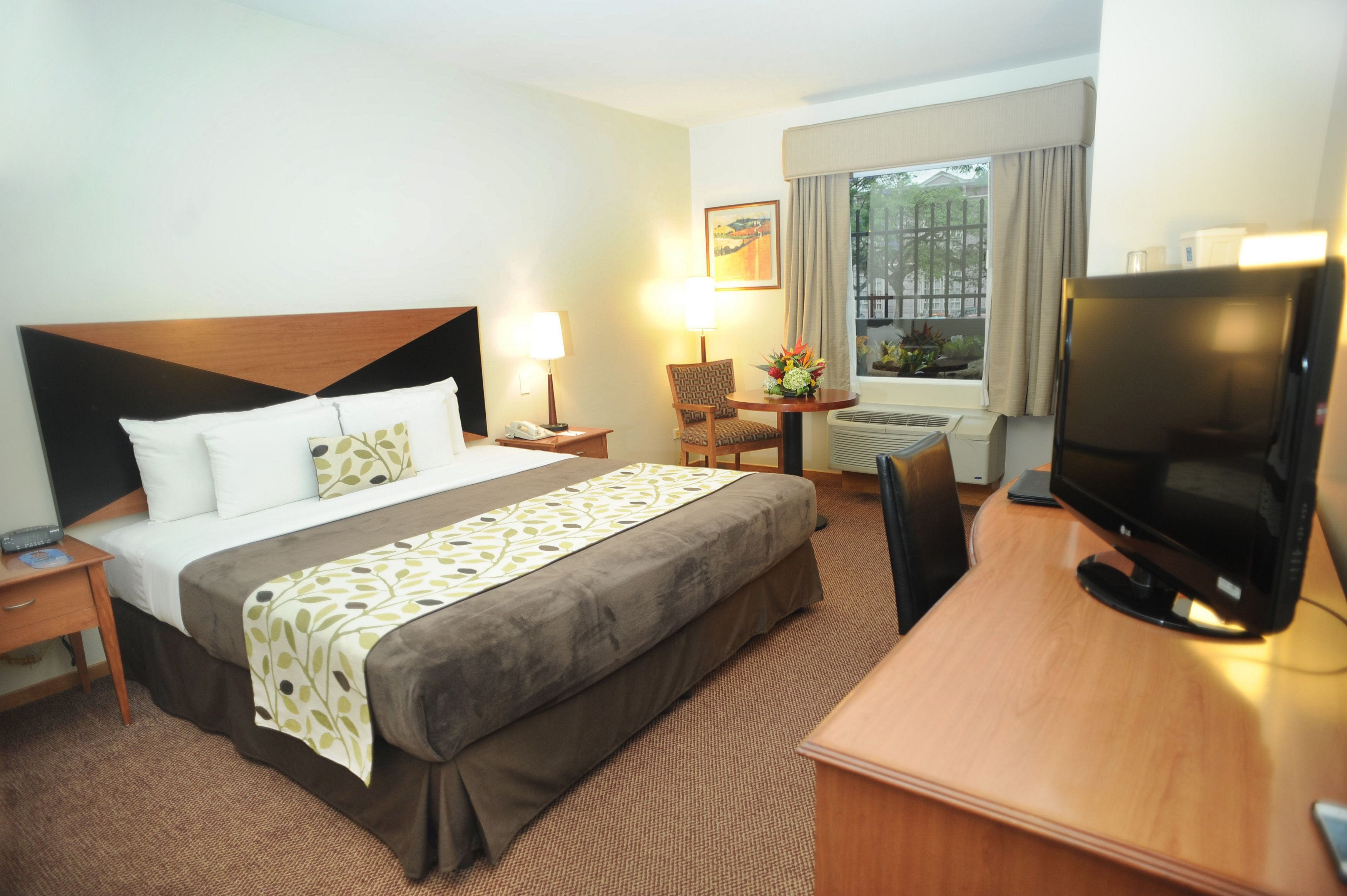 A hotel room with a bed, a desk with a television, a chair, a desk lamp, a small table with a flower arrangement, and a window with curtains.