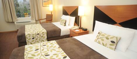 A hotel room with three beds, a window with curtains, a wall-mounted air conditioner, and a nightstand with a lamp.