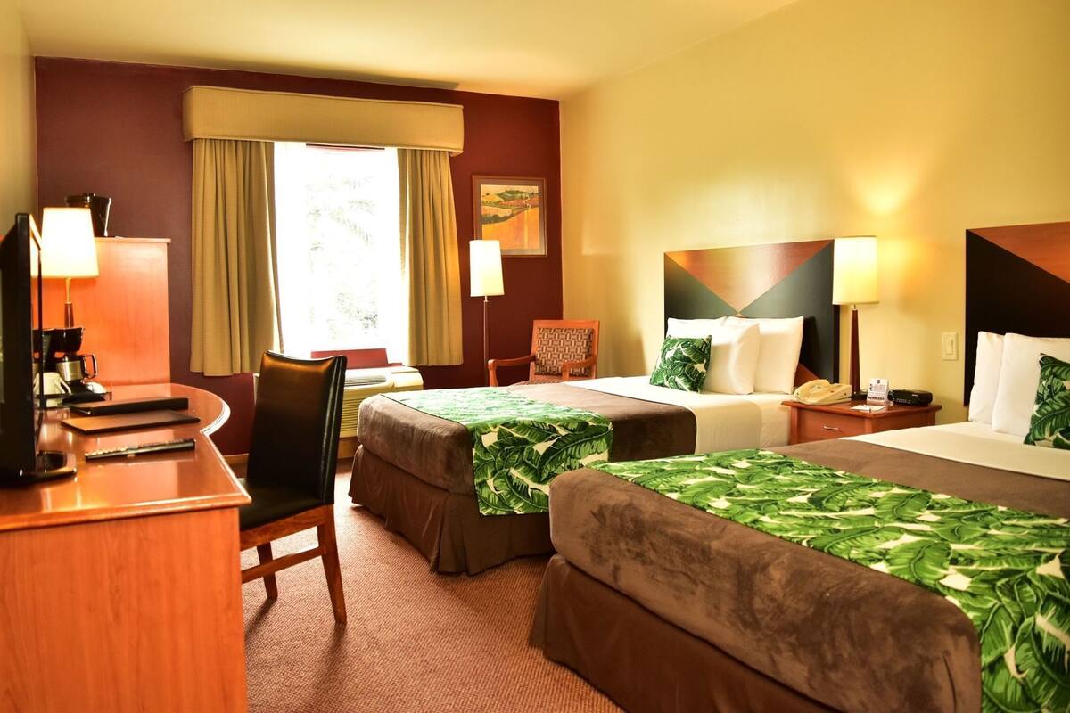 A hotel room with two beds, a desk with a computer, a chair, a lamp, and a painting on the wall.