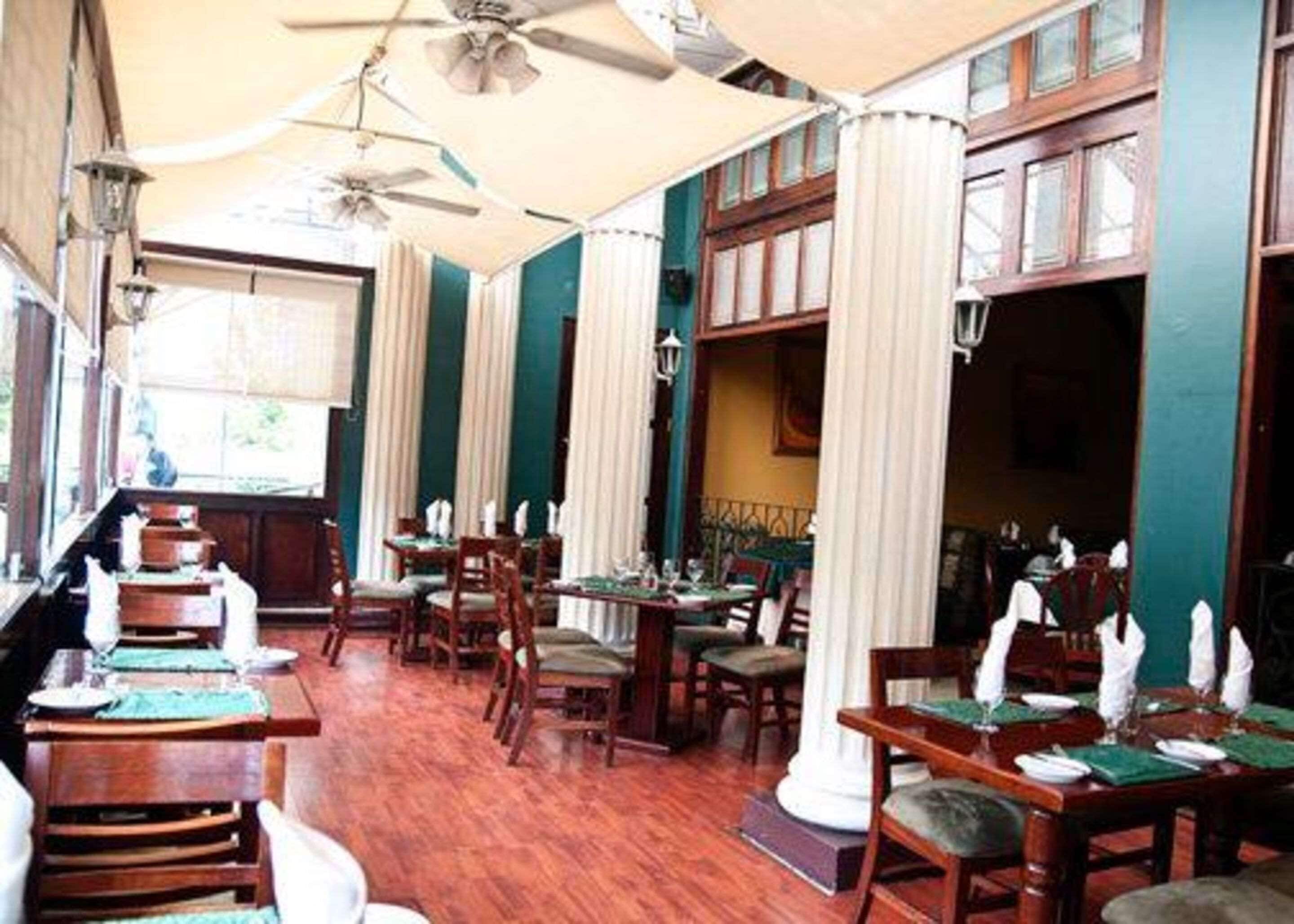 Un restaurant avec des tables et des chaises en bois, des rideaux blancs et un ventilateur de plafond.