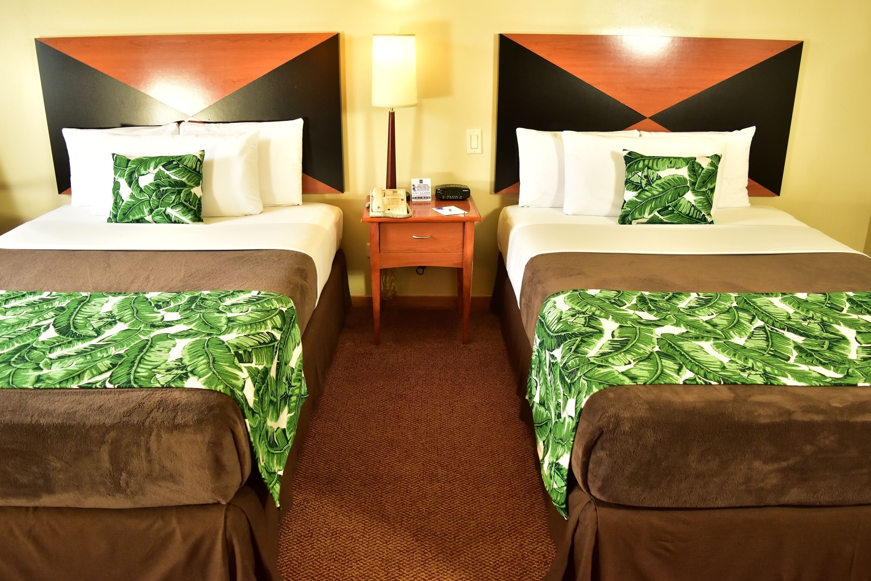 A hotel room with two beds, each with a green leaf-patterned bedspread, a wooden nightstand with a lamp, and a telephone.