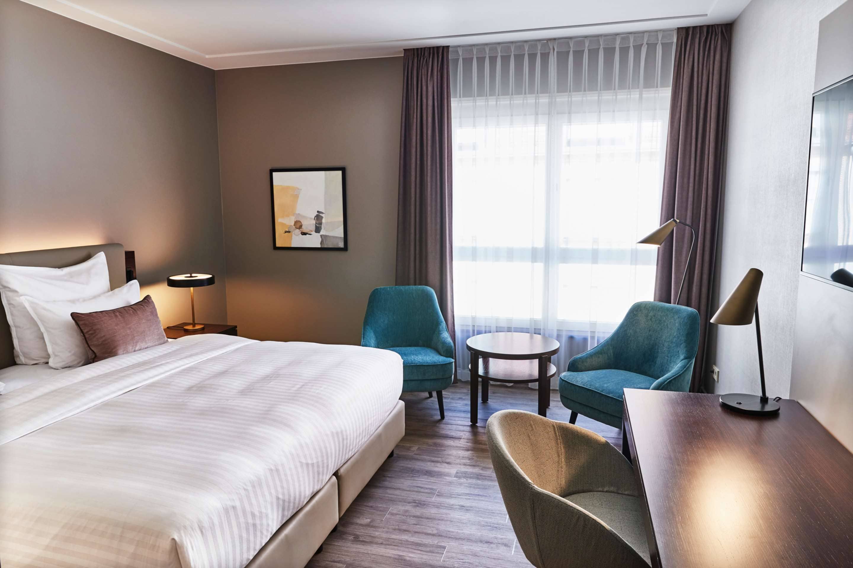 A hotel room with a large bed, two armchairs, a small round table, a desk, a lamp, a framed picture on the wall, and a window with curtains.