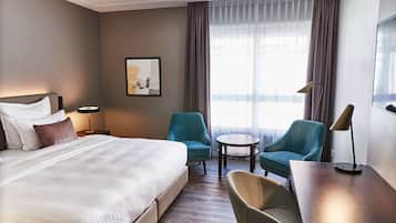 A hotel room with a large bed, two armchairs, a small round table, a desk, a lamp, a framed picture on the wall, and a window with curtains.