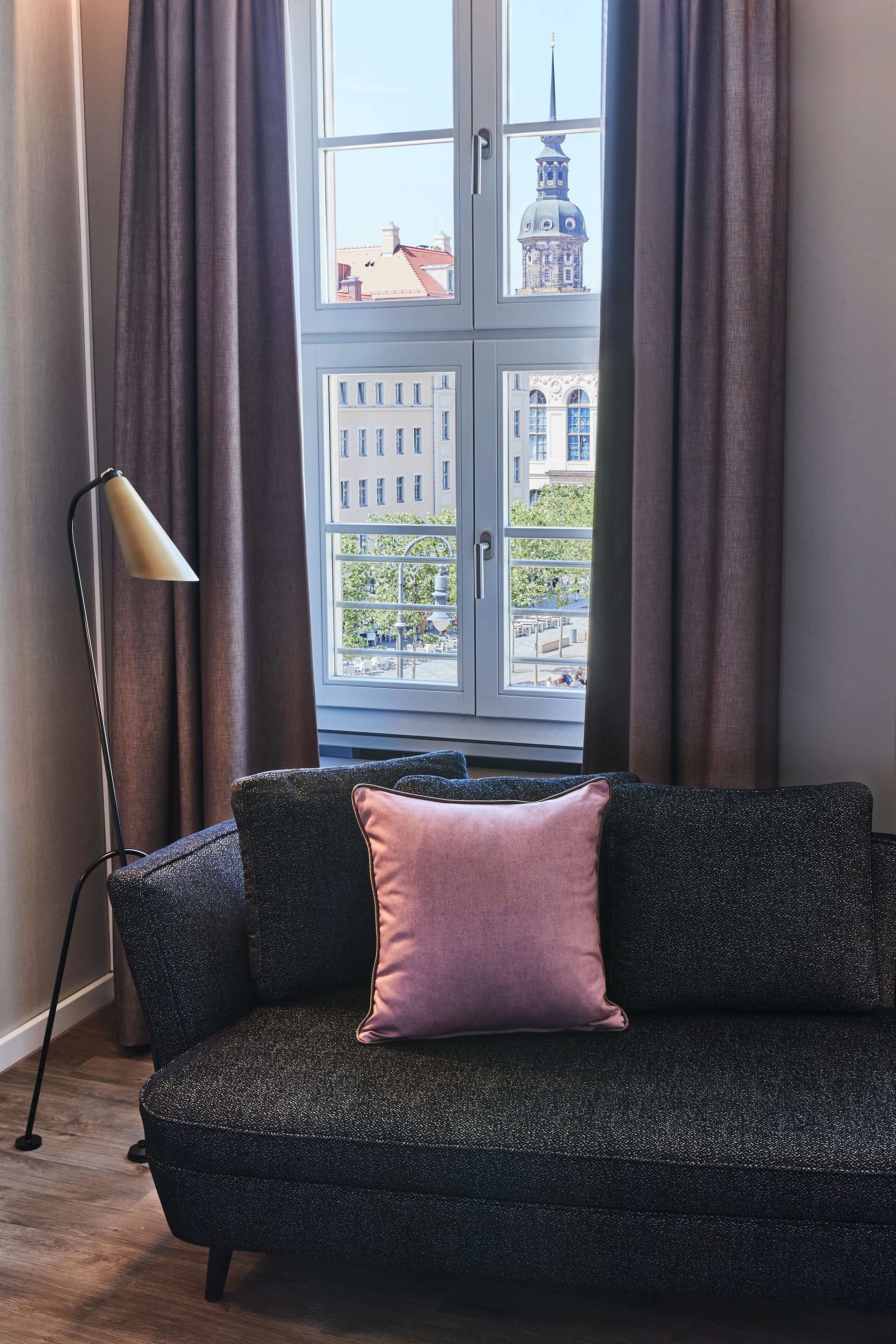 Un canapé à la texture sombre, agrémenté d’un coussin rose, une lampe sur pied et une fenêtre avec des rideaux.