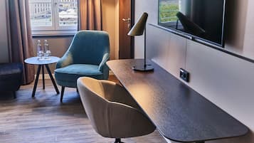 Una habitación de hotel moderna con un televisor grande, un escritorio, un sillón azul, un sillón beige, una pequeña mesa redonda con vasos, y una lámpara.