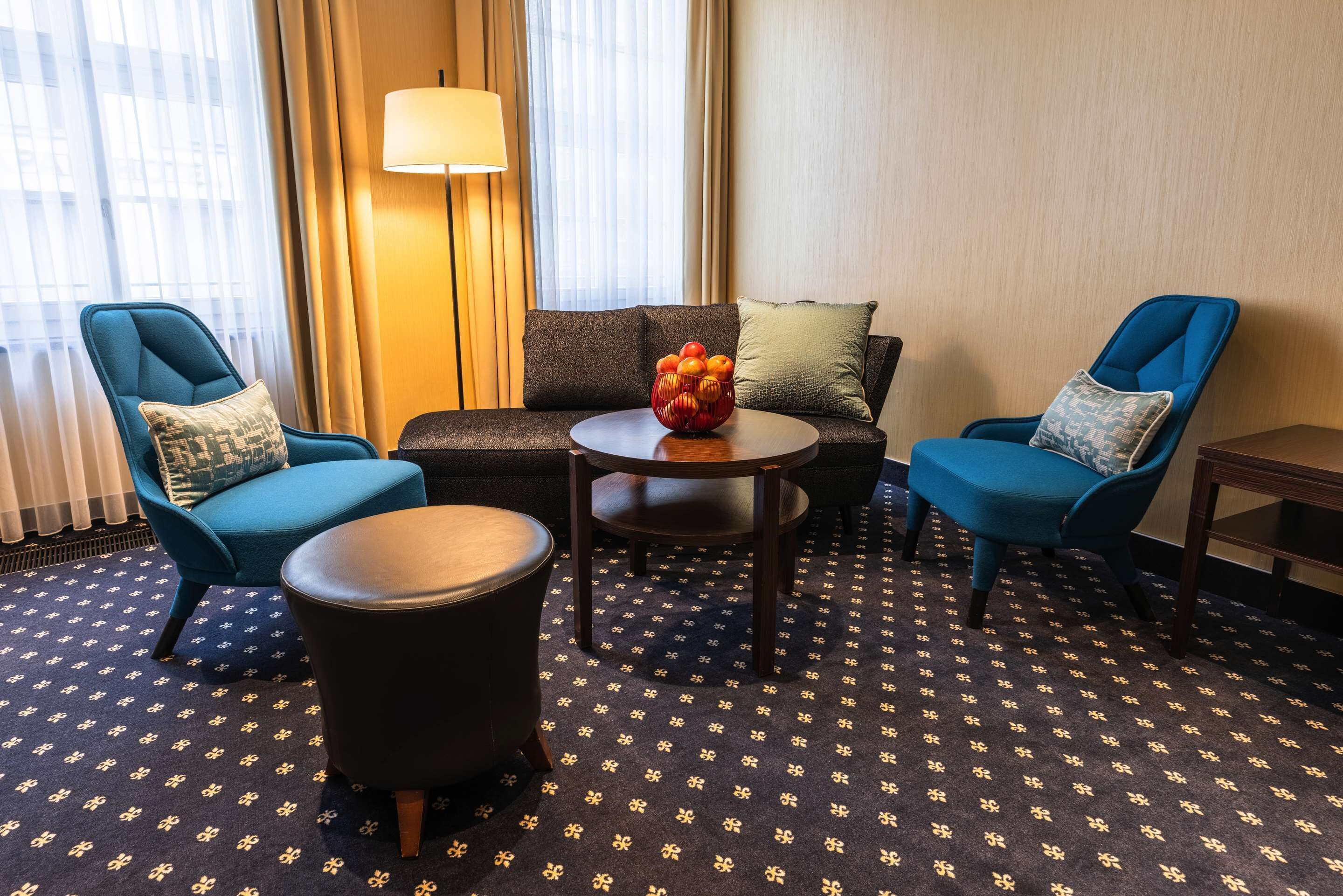 A hotel room with a sofa, two armchairs, a small table with a fruit bowl, a lamp, and patterned carpet.