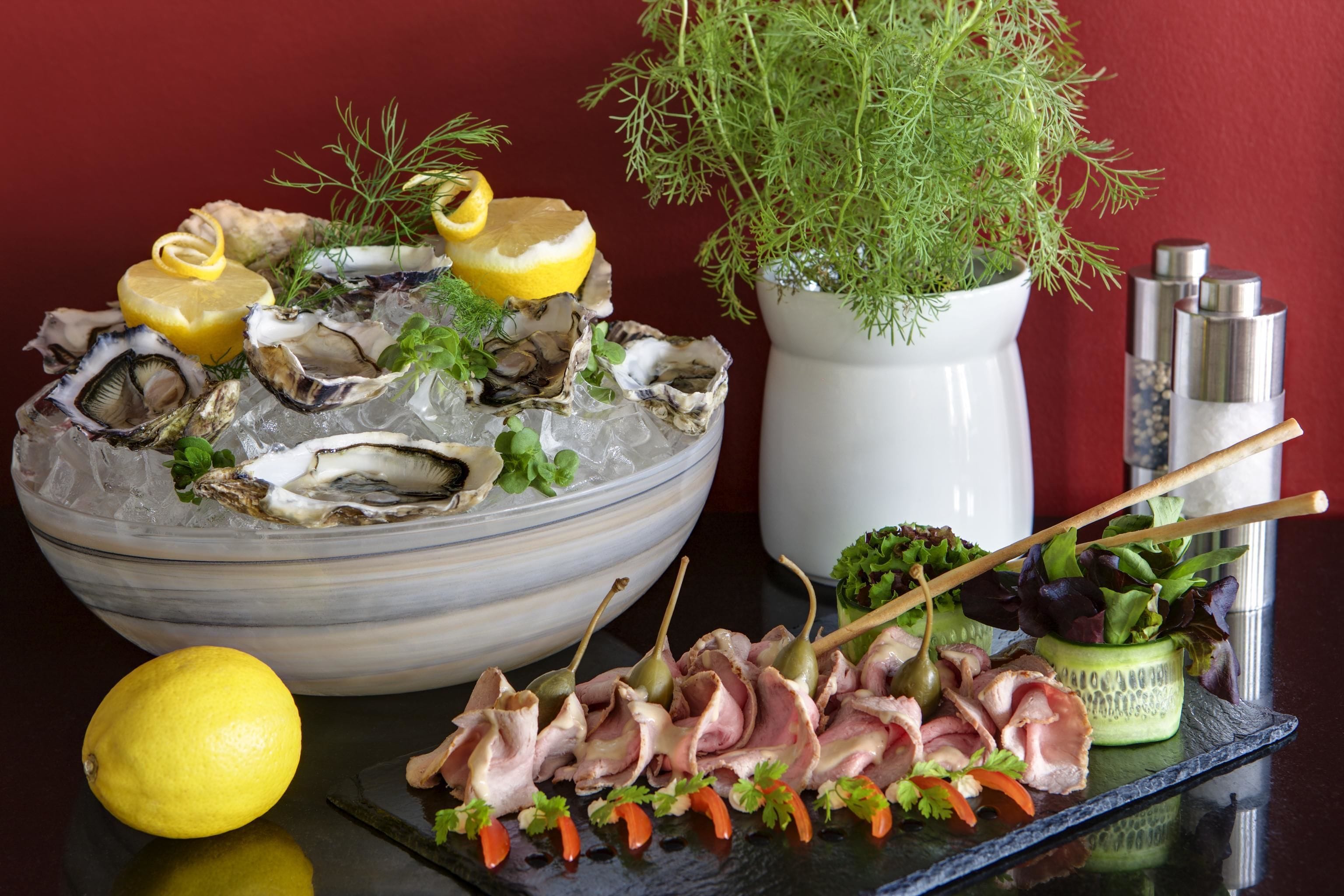 Un bol de ostras sobre hielo, con rodajas de limón y hierbas, una planta en maceta y una bandeja de brochetas de carne con verduras.