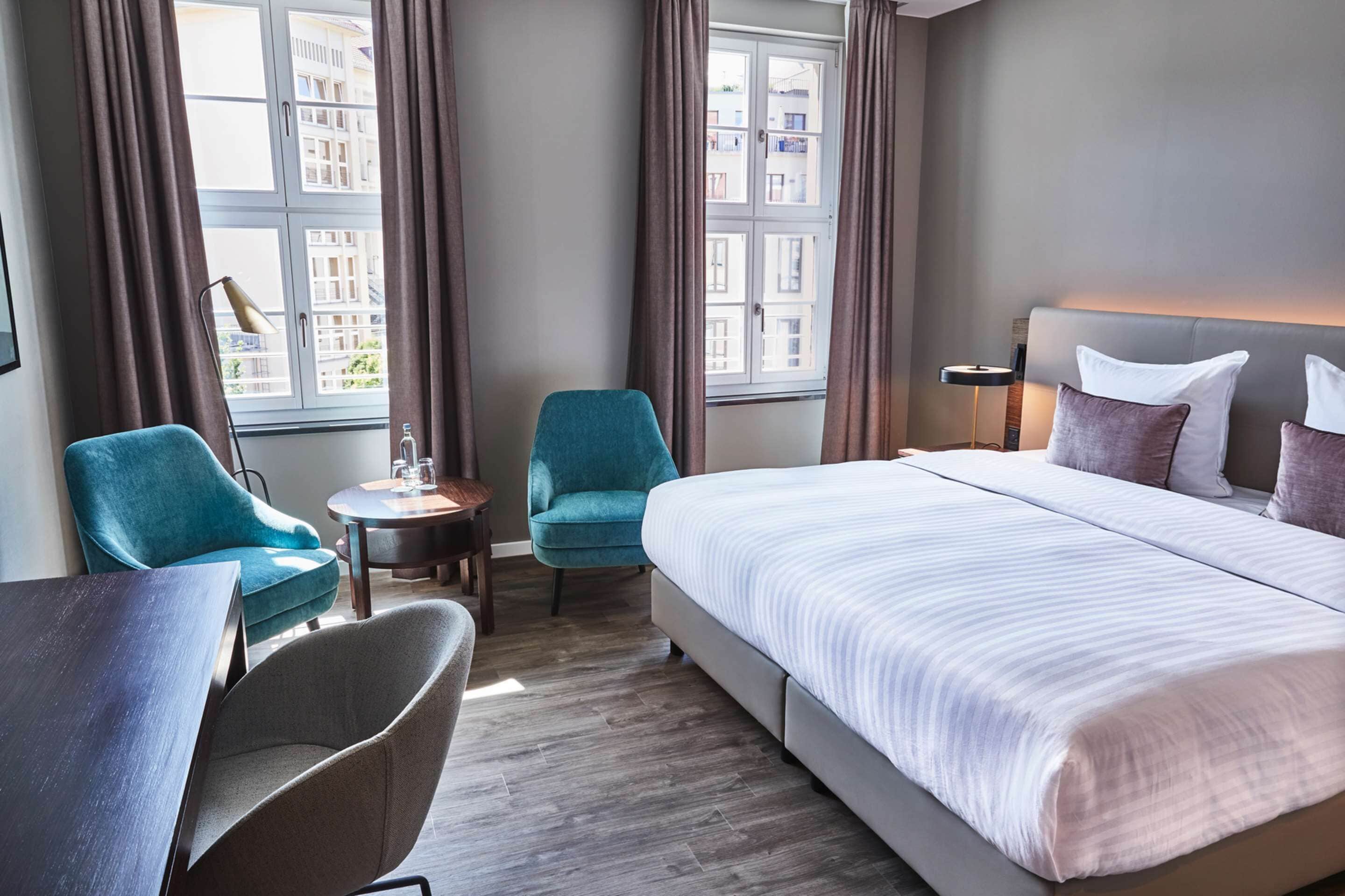 A hotel room with a large bed, two teal armchairs, a small round table, and a lamp.