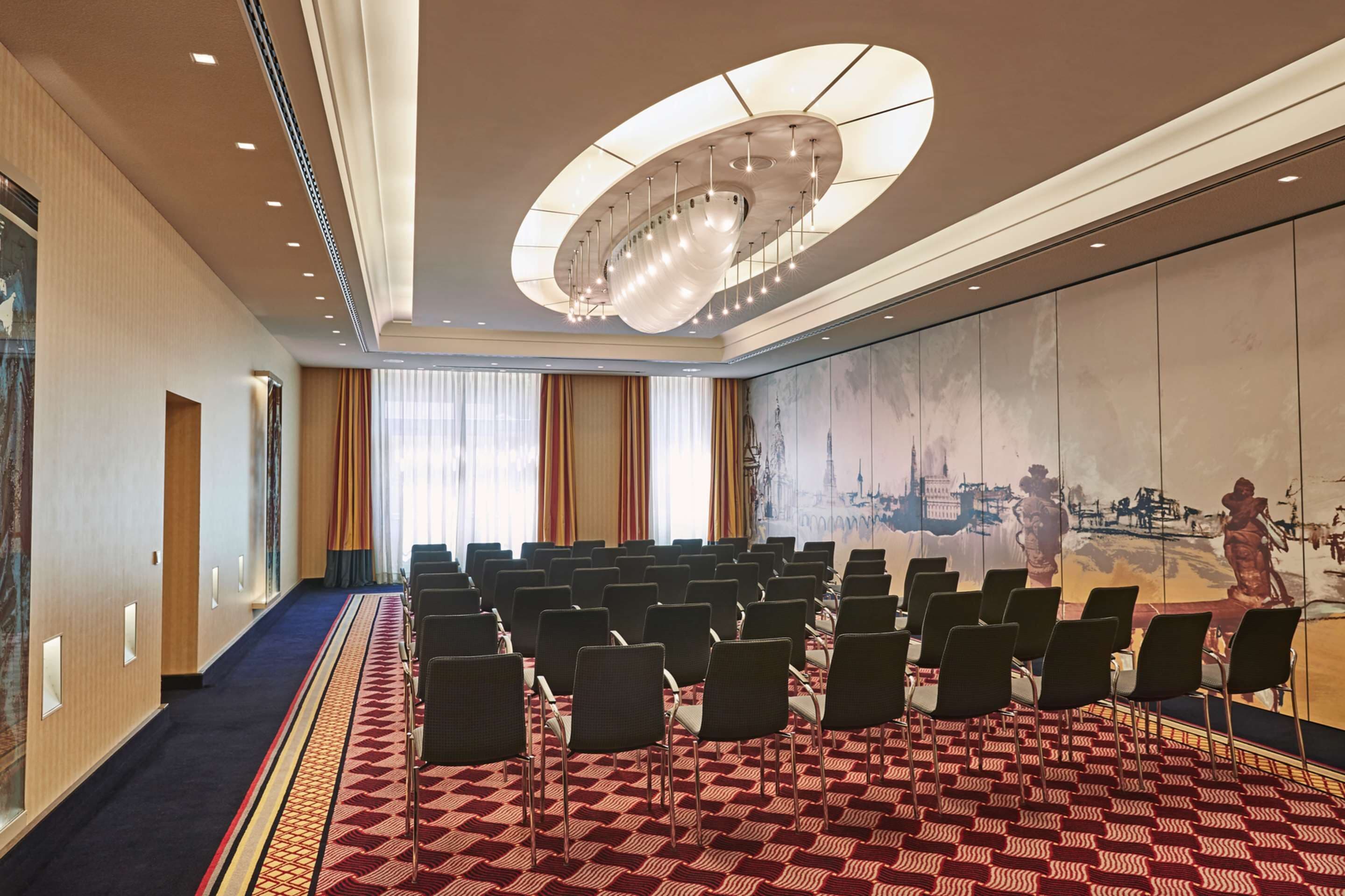 A conference room with rows of chairs, a patterned carpet, a large mural on the wall, and a modern ceiling light fixture.