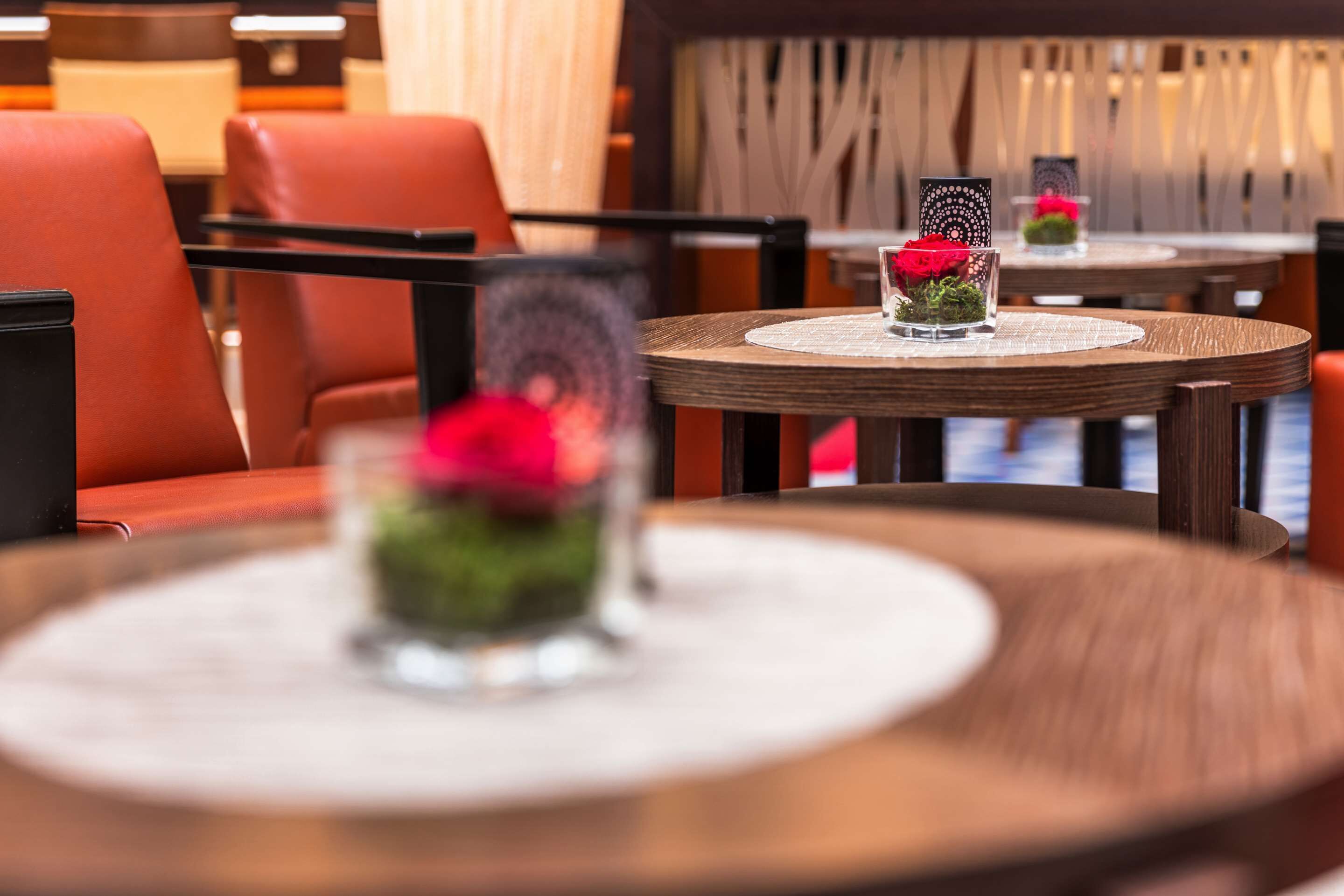 A restaurant setting with round tables, red leather chairs, and a decorative vase with flowers on each table.