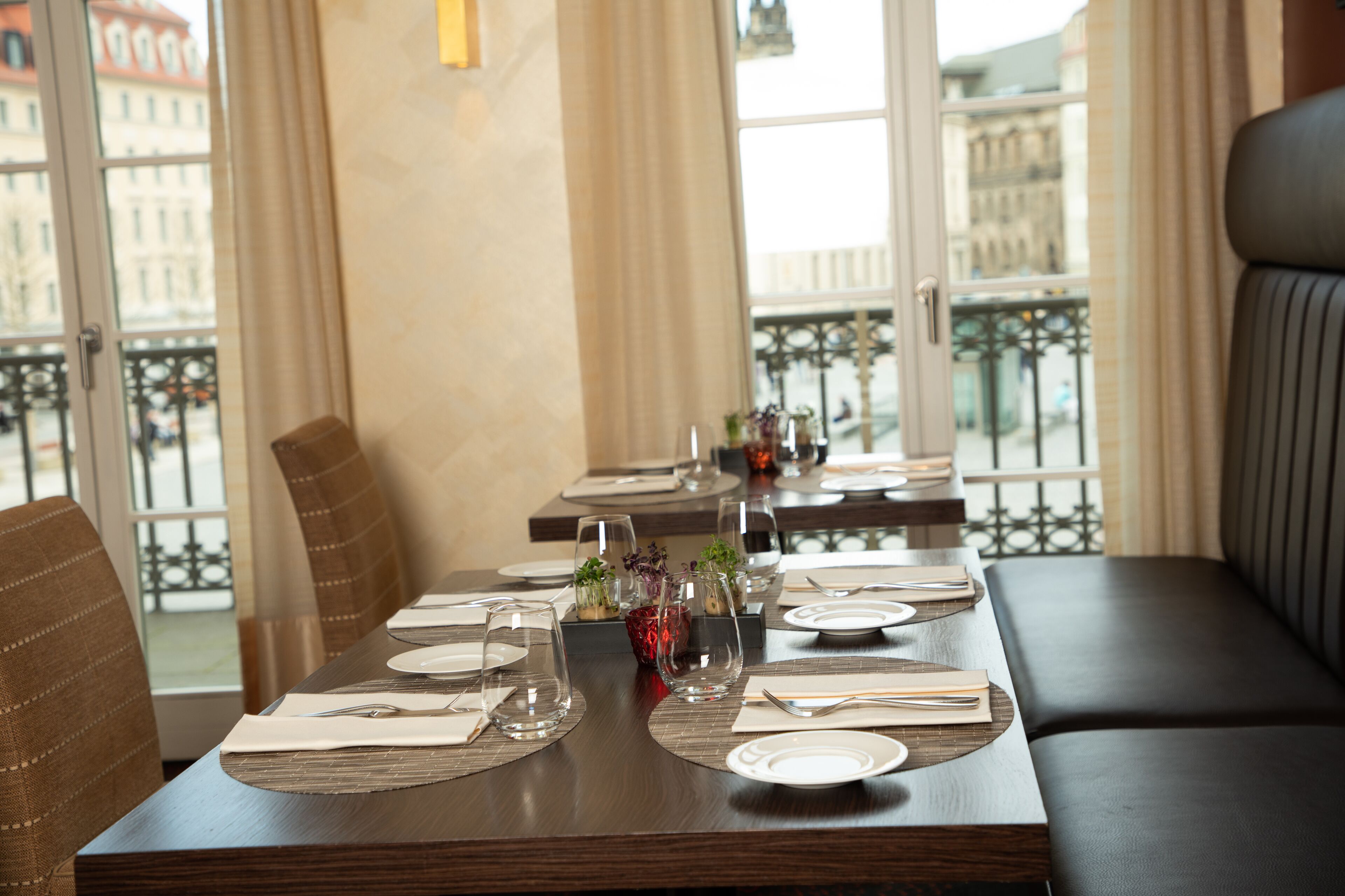 A dining area with tables set for two, featuring a view of the city through large windows.