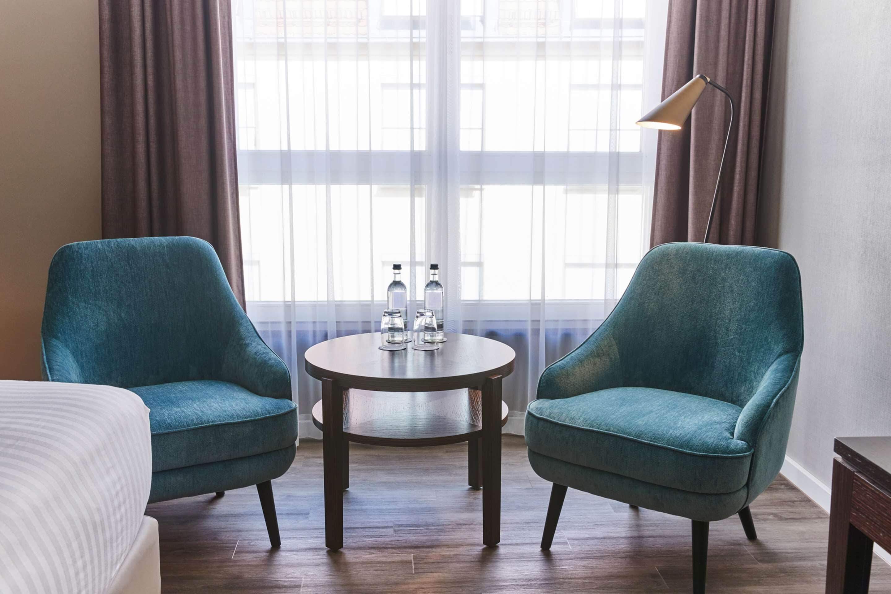Ein Hotelzimmer mit zwei türkisfarbenen Sesseln, einem kleinen runden Tisch mit Wasserflaschen und Gläsern, einer Lampe und einem Bett.