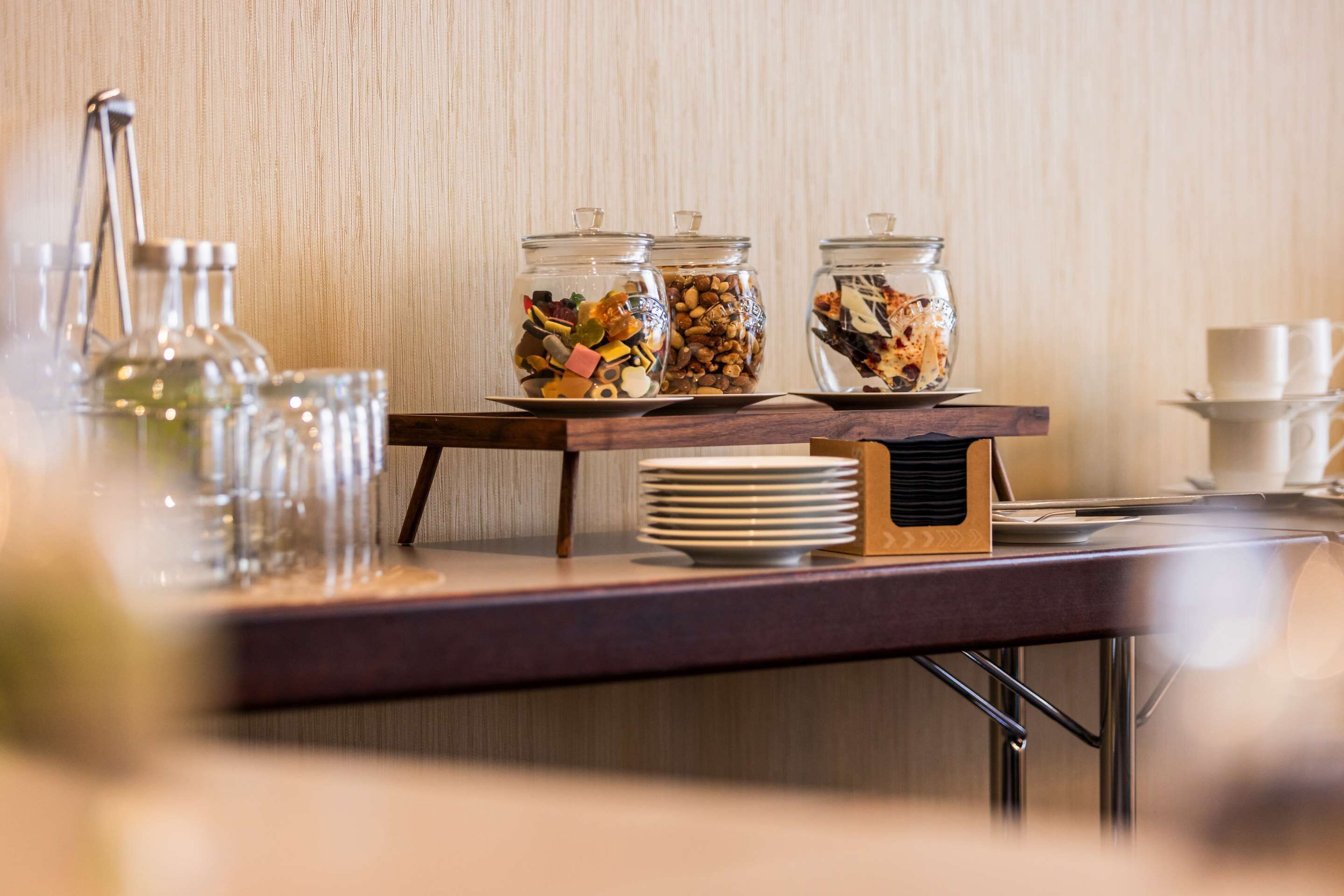 A buffet setup with glass jars containing various food items, a stack of white plates, and a coffee station with cups and a thermos.