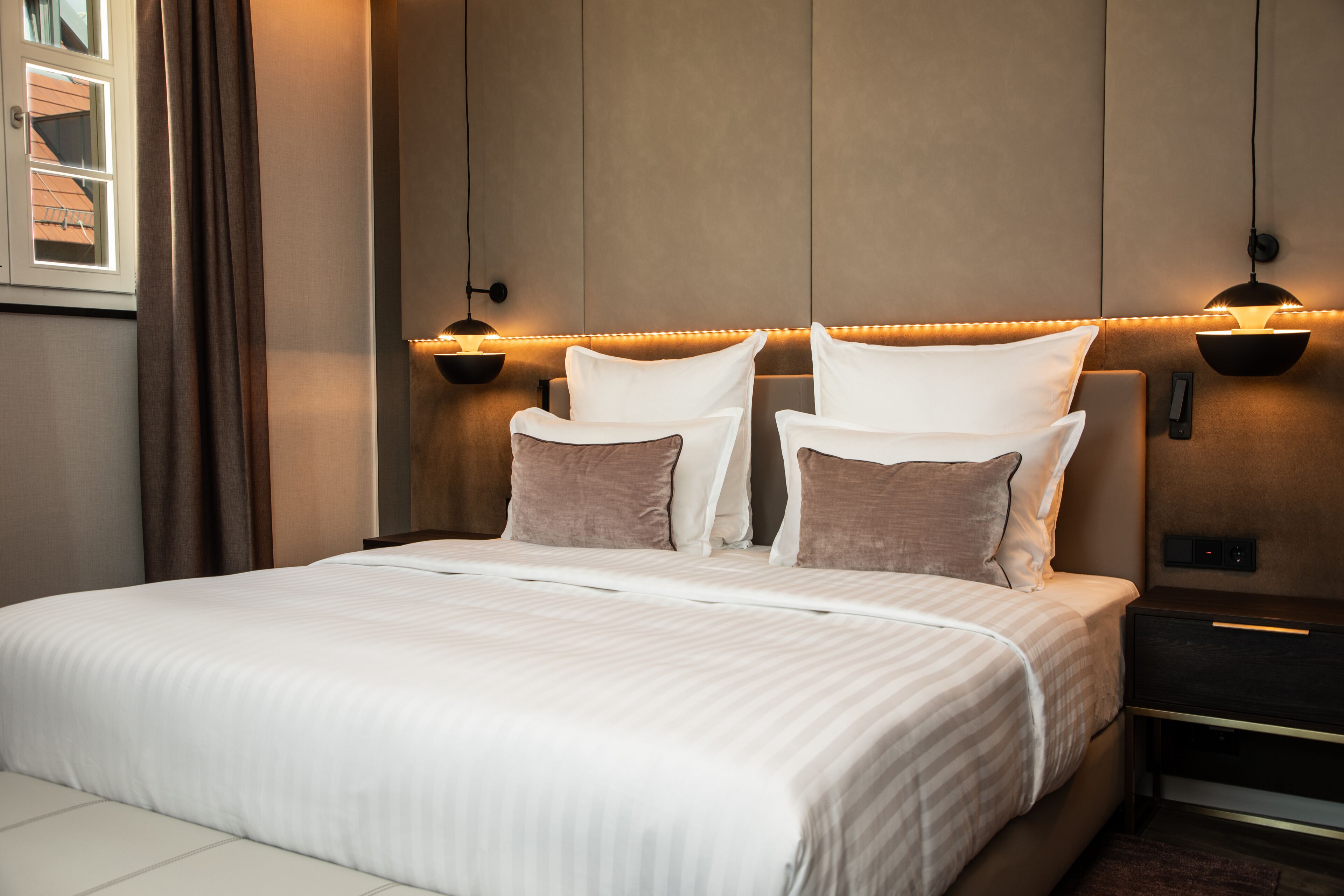A neatly made bed with white linens and two pillows, a bedside table with a lamp, and a wall-mounted light fixture.
