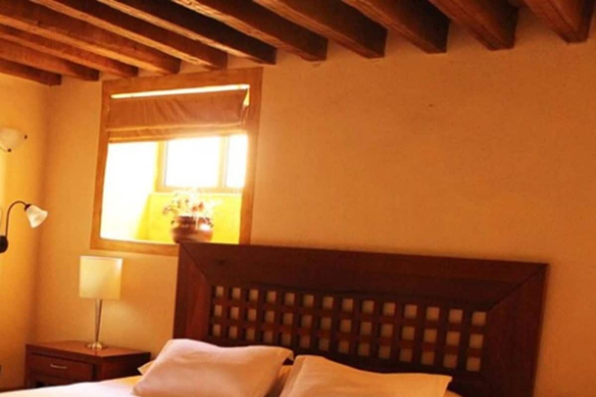 A bedroom with a wooden headboard, a bed with white linens, a nightstand with a lamp, and a window with blinds.