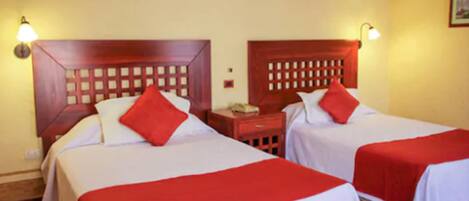 A hotel room with two beds, each with a wooden headboard and red pillows. There are bedside tables with lamps and a small nightstand with a phone.