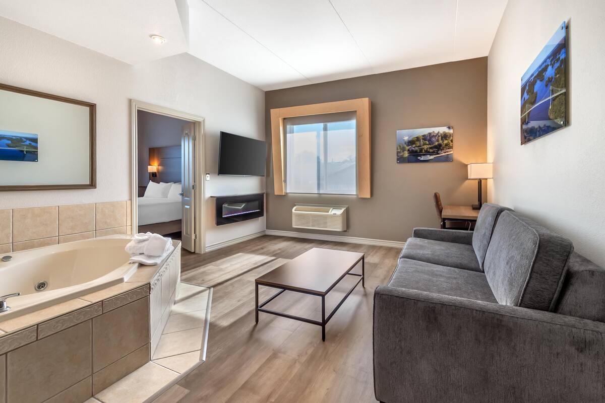 A modern hotel room with a bathtub, a gray sofa, a small table, and a large window.