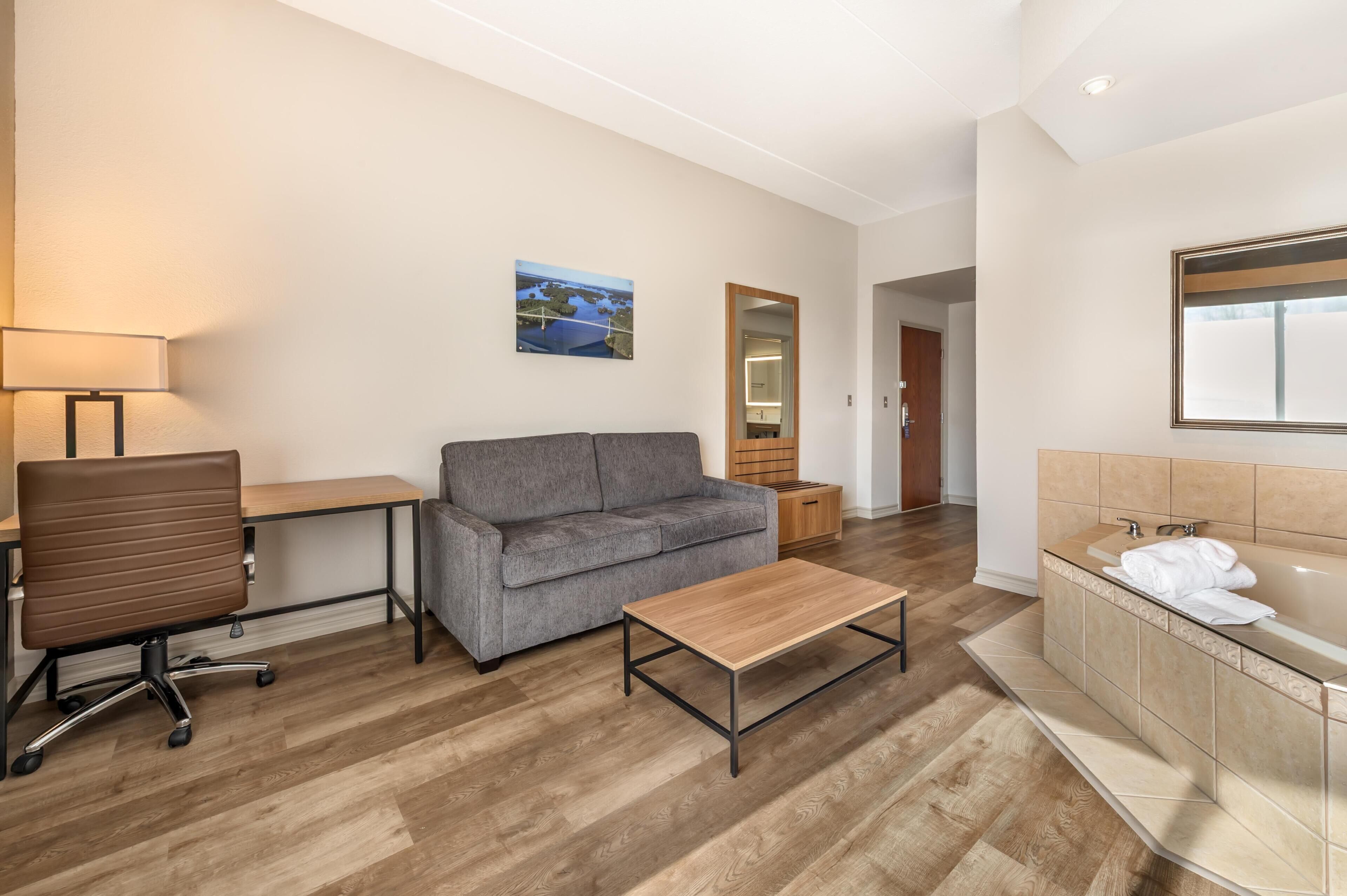 Une chambre d’hôtel avec un plancher en bois, un canapé gris, une table basse en bois, un bureau avec une lampe, un grand miroir et une salle de bain avec une baignoire et un lavabo.