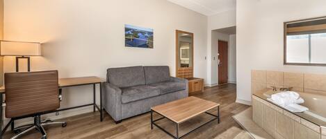 Une chambre d’hôtel avec un plancher en bois, un canapé gris, une table basse en bois, un bureau avec une lampe, un grand miroir et une salle de bain avec une baignoire et un lavabo.