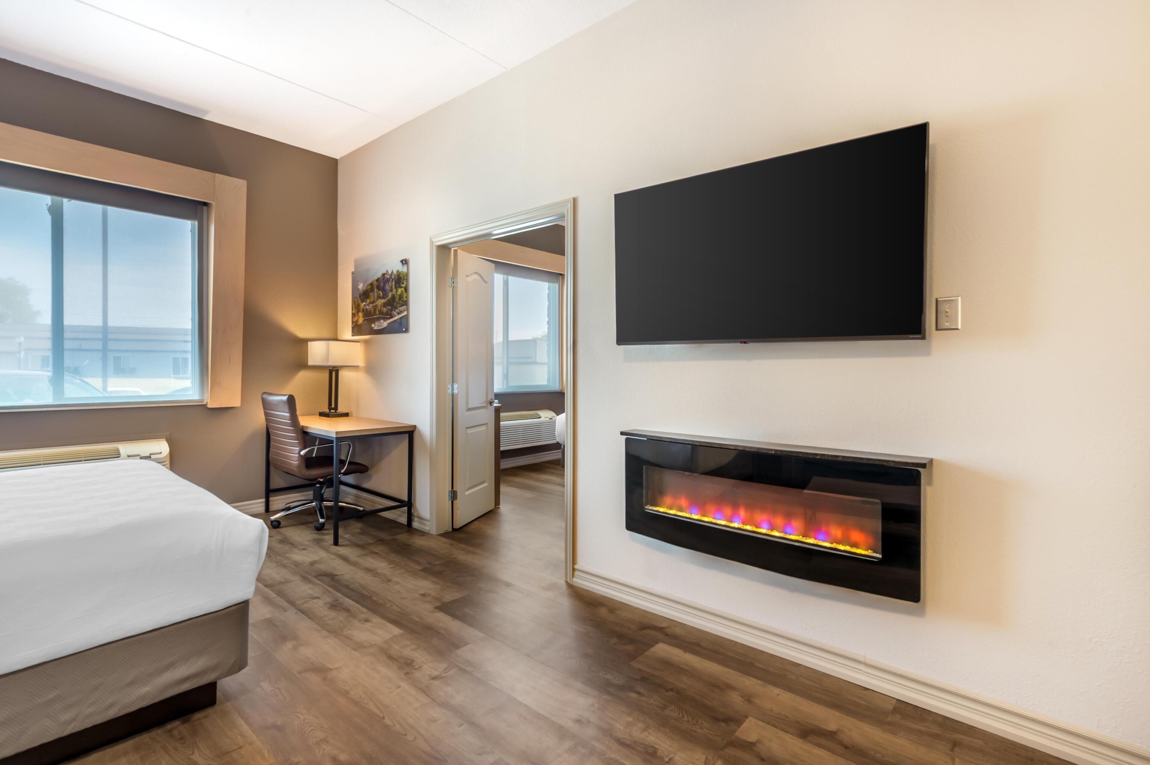 A hotel room with a bed, desk, chair, TV, and a fireplace.